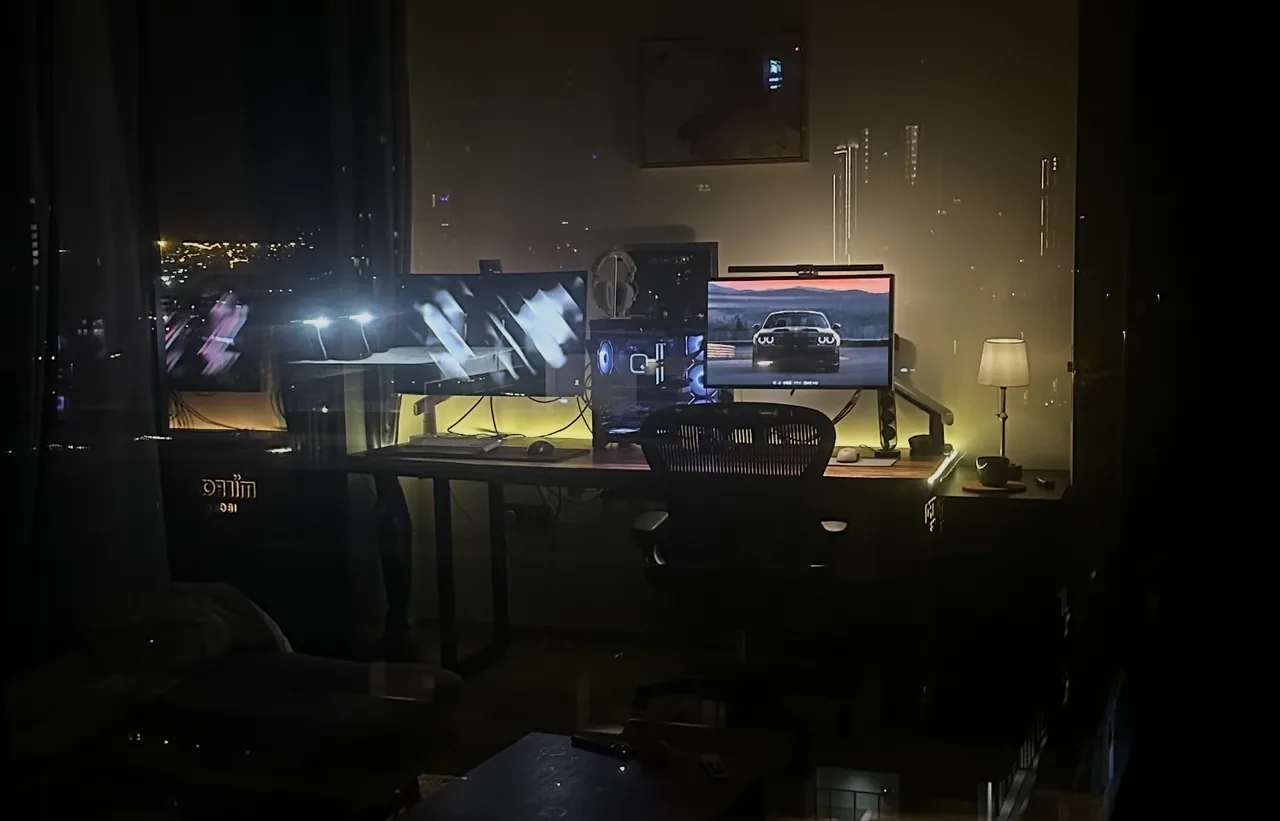Desk setup at night with two monitors and warm lighting, seen through a window